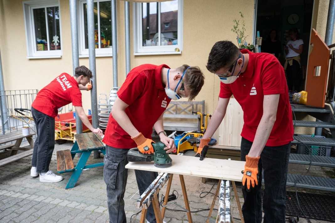 drei Männer in roten T-Shirts arbeiten mit Holz vor einem Haus