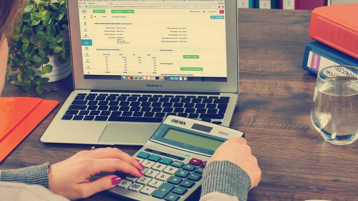 Person sitzt vor Laptop und hat Taschenrechner in der Hand