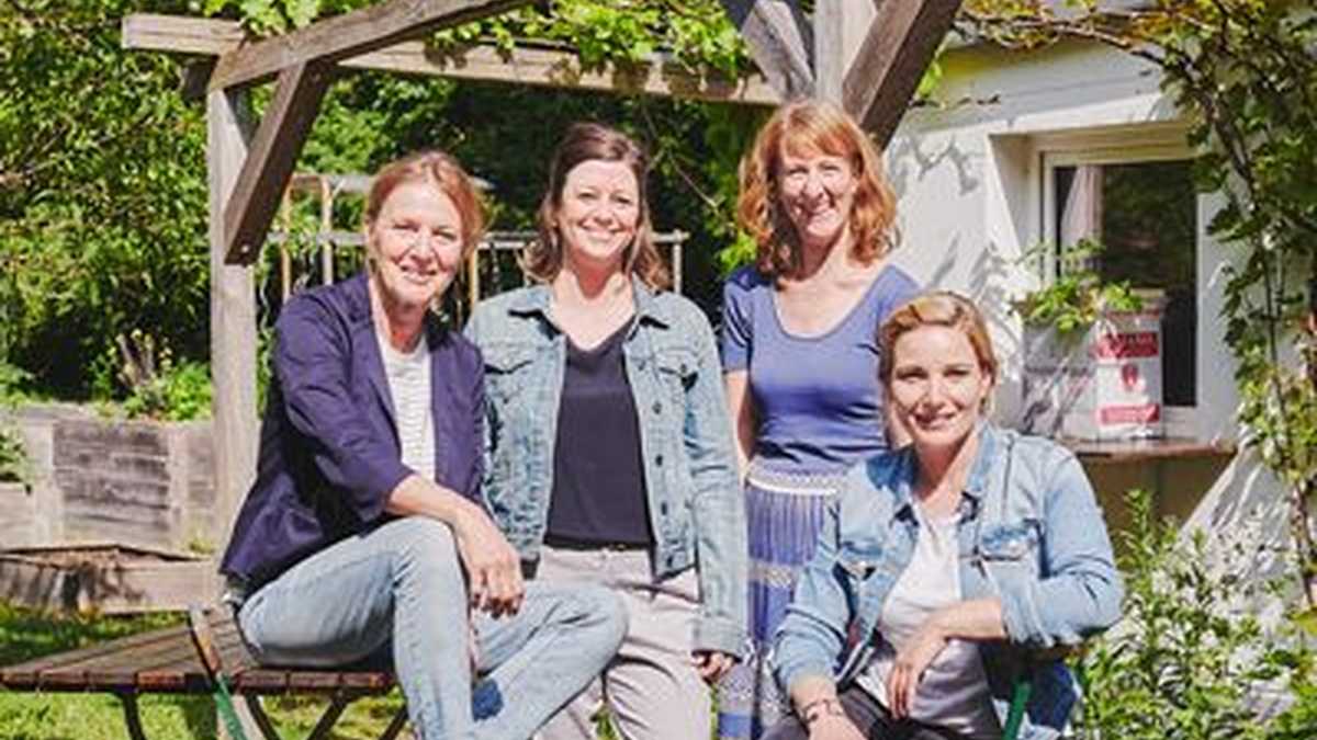 vier Frauen mit blauen Oberteilen sitzen auf oder stehen um Gartenmöbel